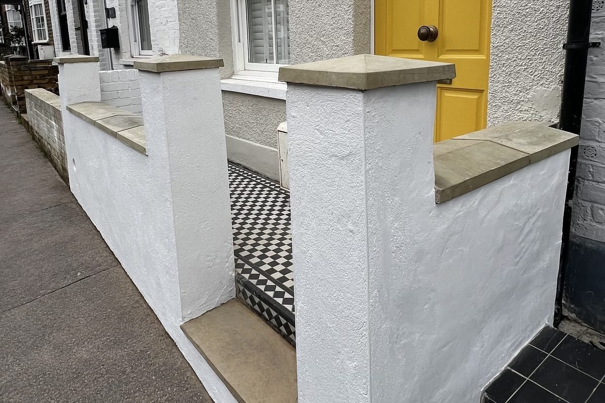 A finished front garden, white rendered walls with stone copings, a black and white tiled path and a yellow front door