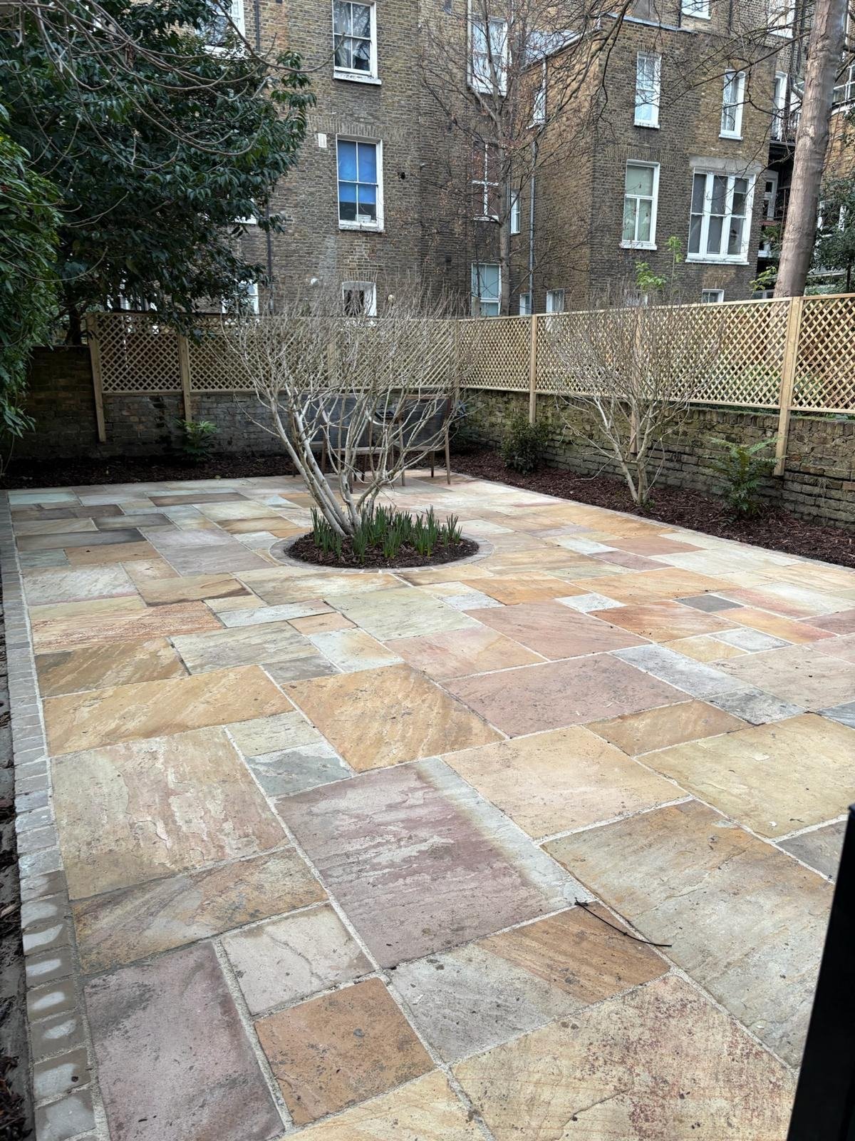 The same garden finished, sandstone terrace with a circular planting bed around a tree and new trellis on the wall