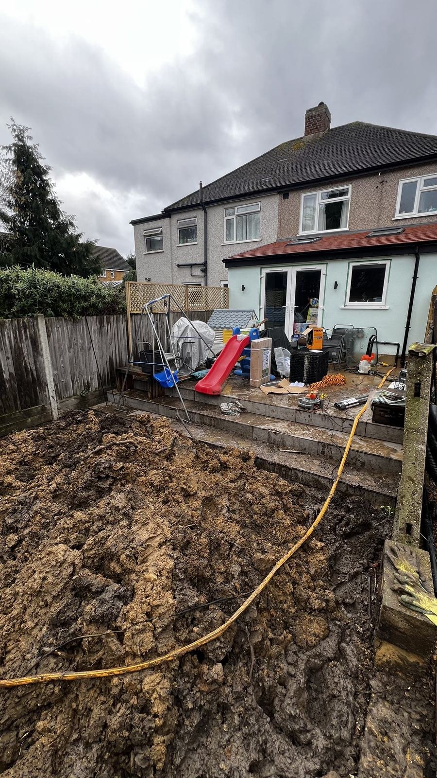Rear garden mid-works, excavated earth and old fencing beside a semi-detached house